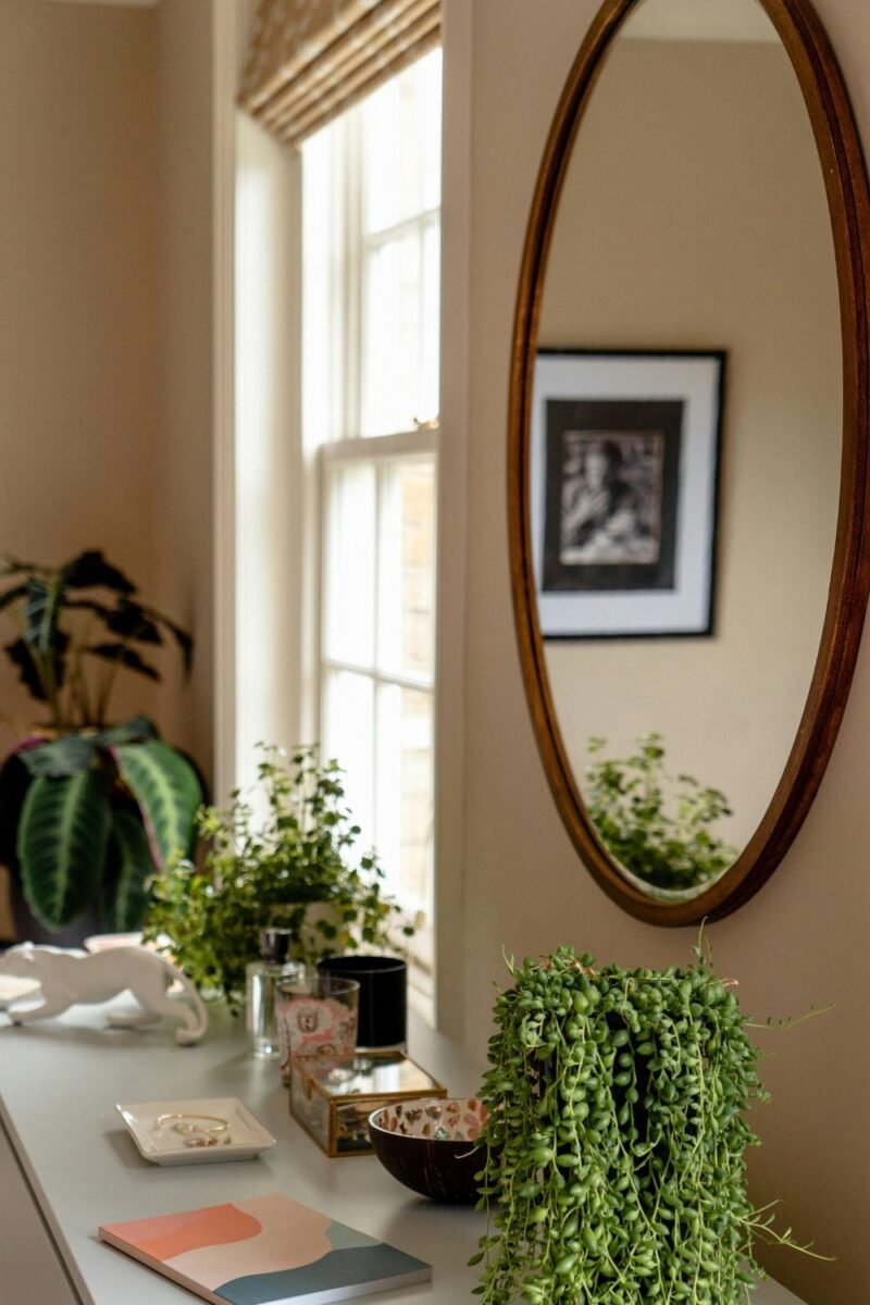 Oval mirror hung beside a bedroom window, bouncing daylight back into the room next to trailing plants