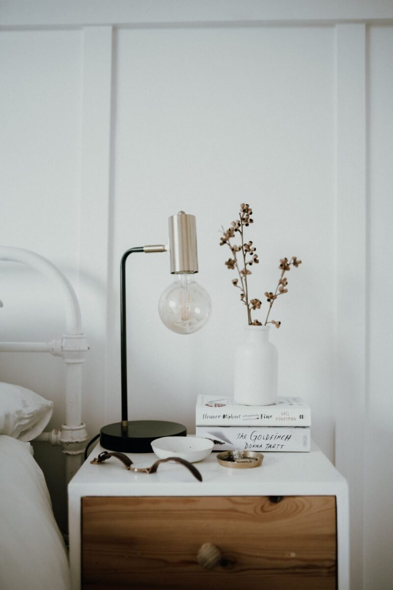 Styled nightstand with a black lamp, stacked books and a small vase of dried stems beside the bed