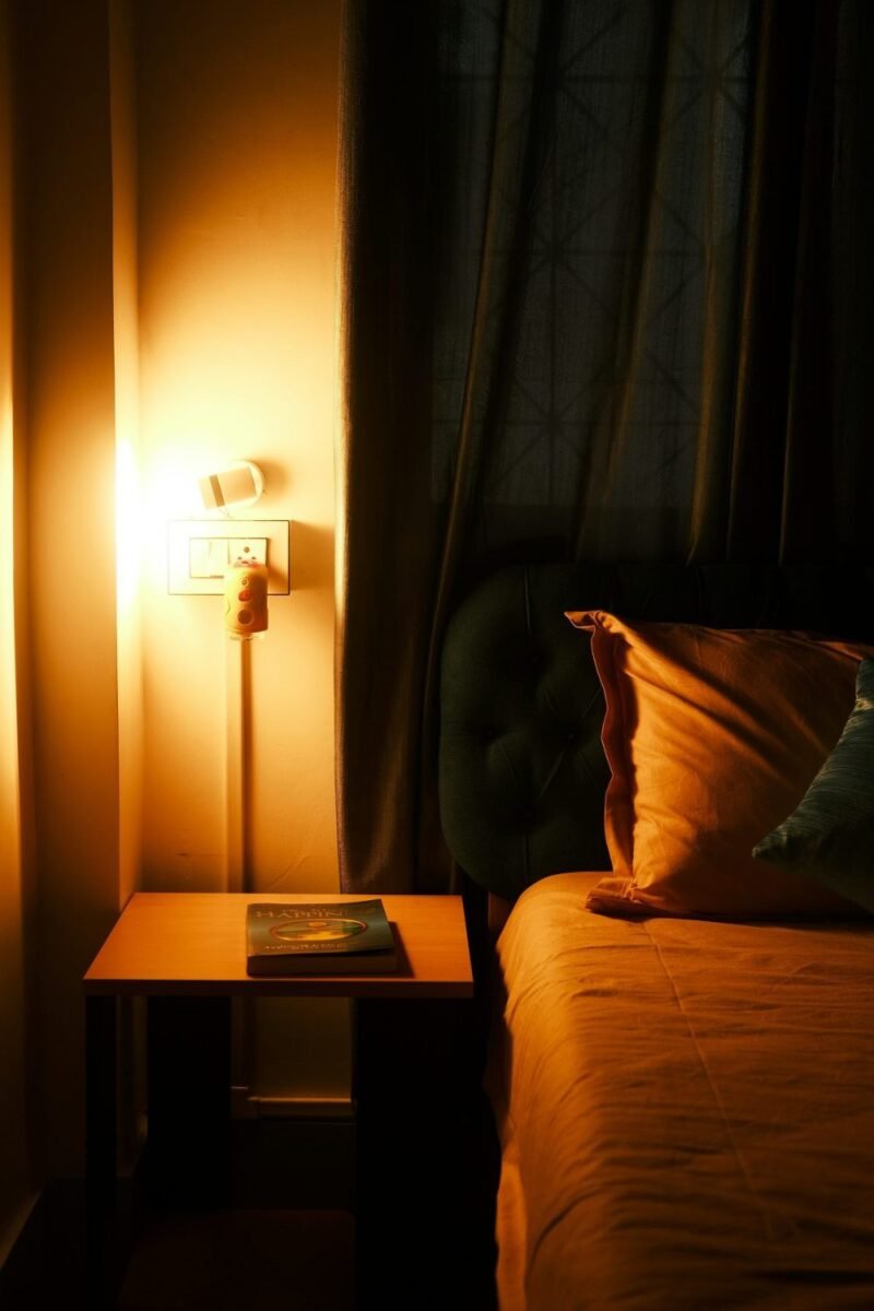 Small bedroom at night with a warm wall lamp glowing over a wooden nightstand and a book