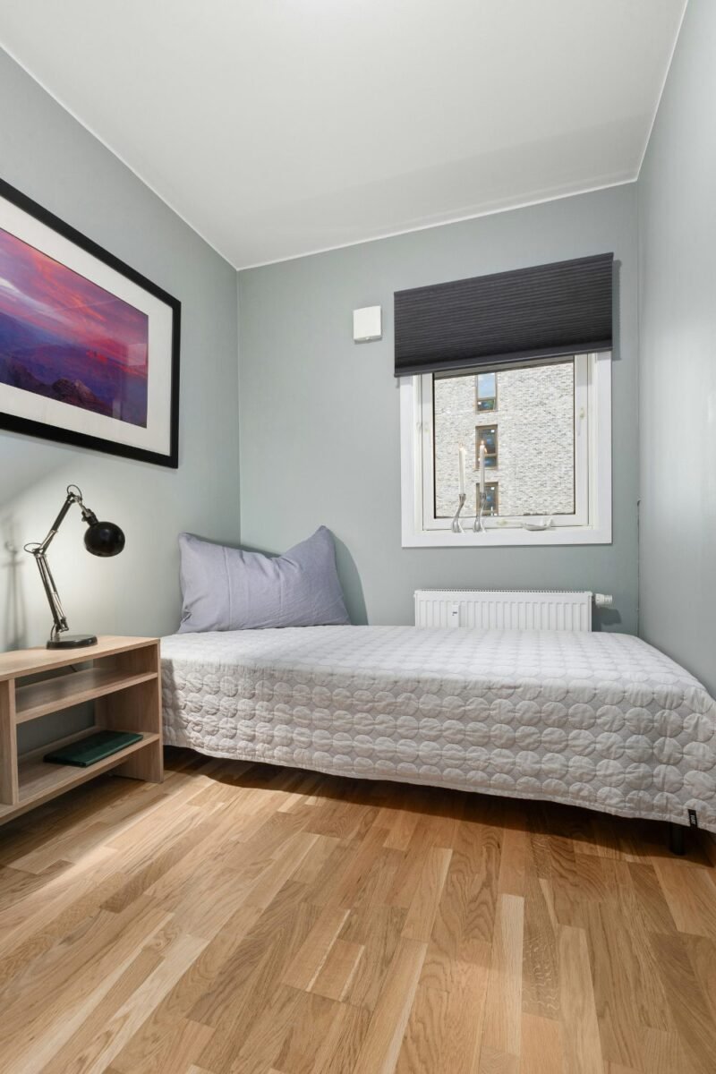 Bed pushed against the wall of a small apartment bedroom, leaving a wide stretch of bare wood floor open