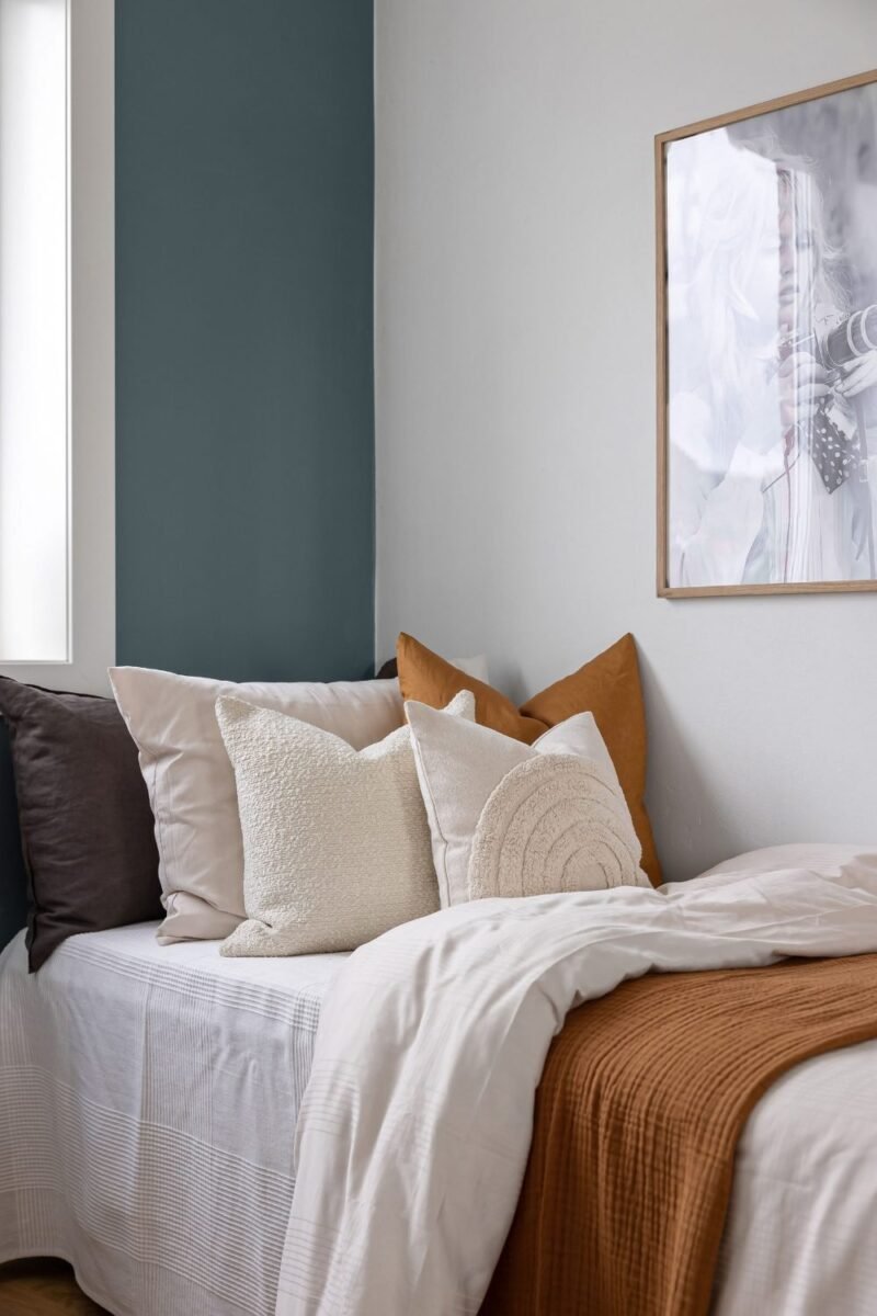 Corner of a bed in a small apartment bedroom layered with a waffle cushion, a knit pillow and a folded rust throw