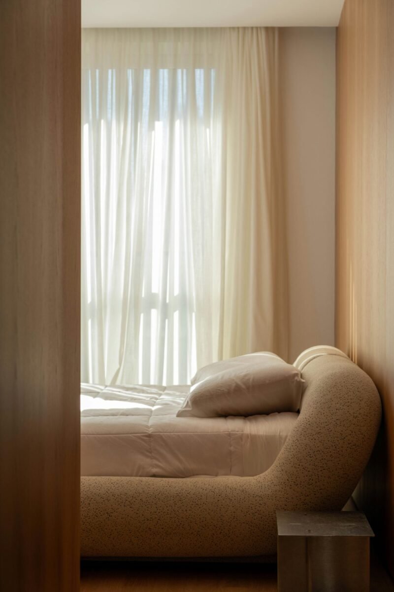 Long sheer curtains hung high beside a window in a small apartment bedroom, with a curved upholstered seat catching the daylight