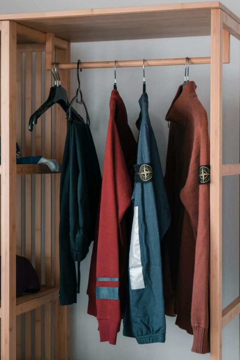 Open wooden clothing rack in a small apartment bedroom, holding hanging jackets above a slatted shelf