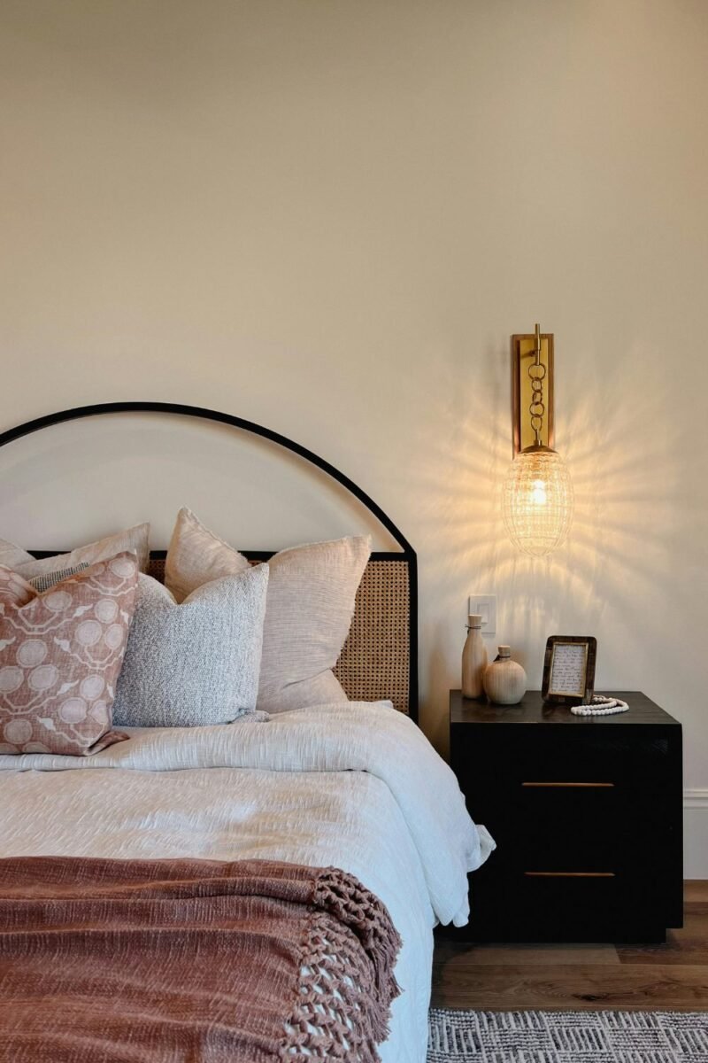 Lit brass wall sconce beside an arched headboard in a small bedroom, freeing up the dark nightstand below