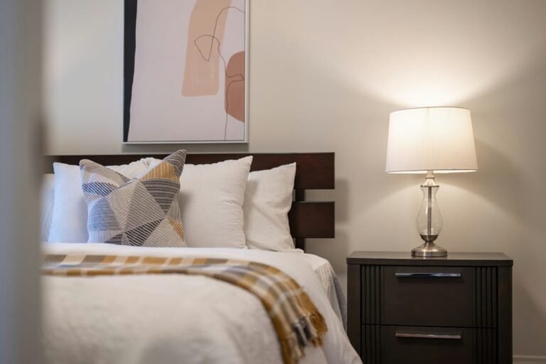 Small bedroom lit by a bedside lamp on a dark nightstand, with white bedding, mustard accents and abstract art above the bed