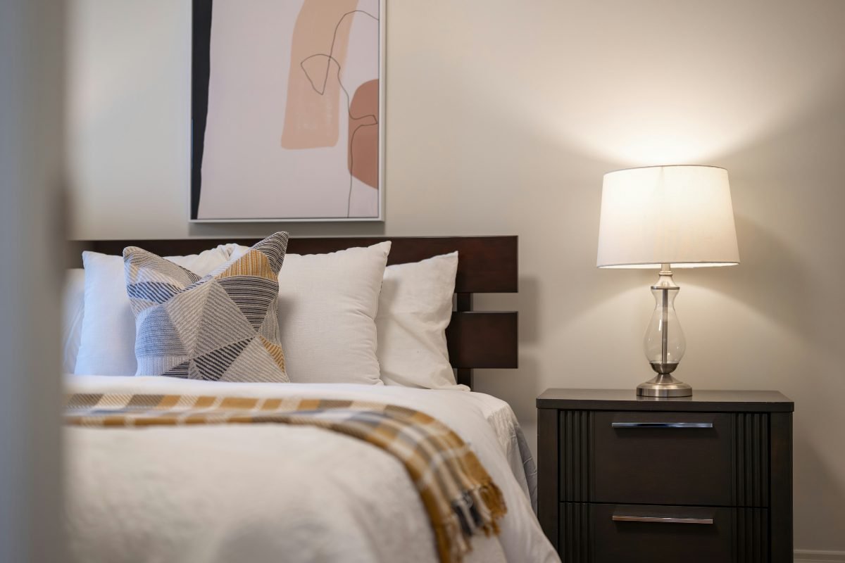 Small bedroom lit by a bedside lamp on a dark nightstand, with white bedding, mustard accents and abstract art above the bed