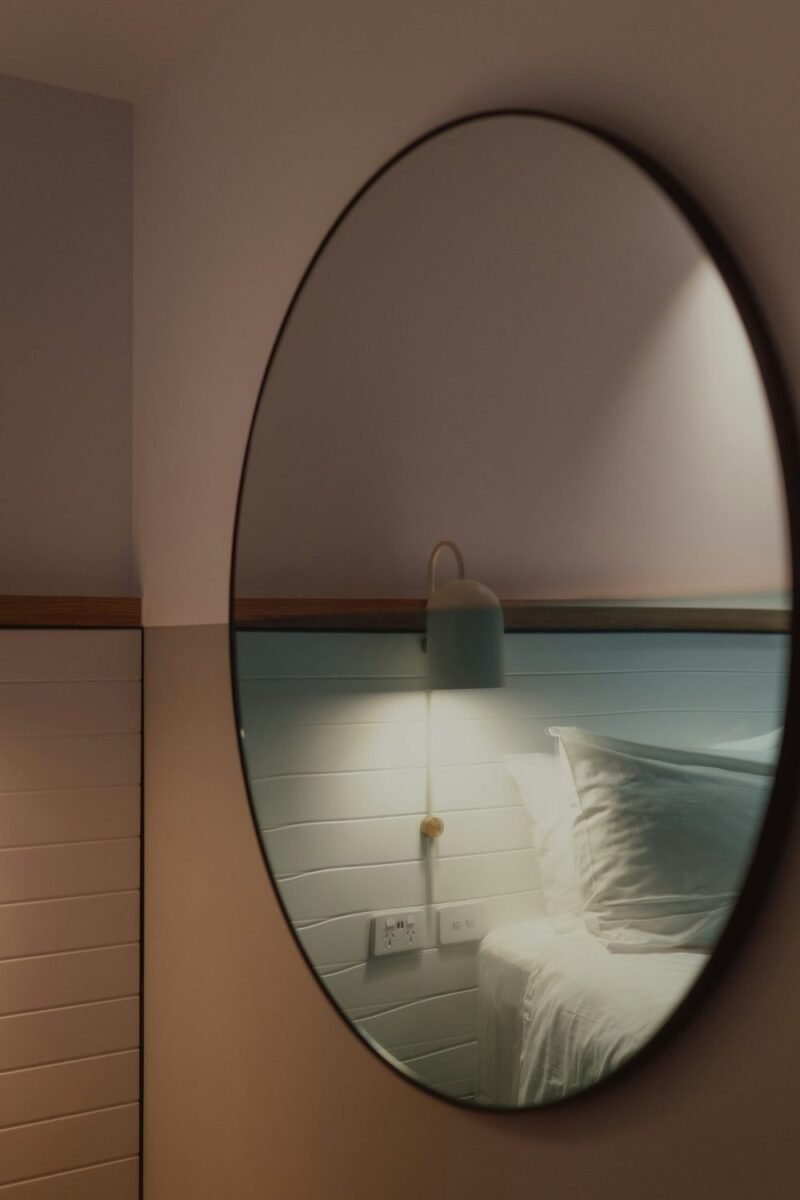 Large oval wall mirror reflecting the bed and a wall light, bouncing light around a small bedroom to make it feel larger