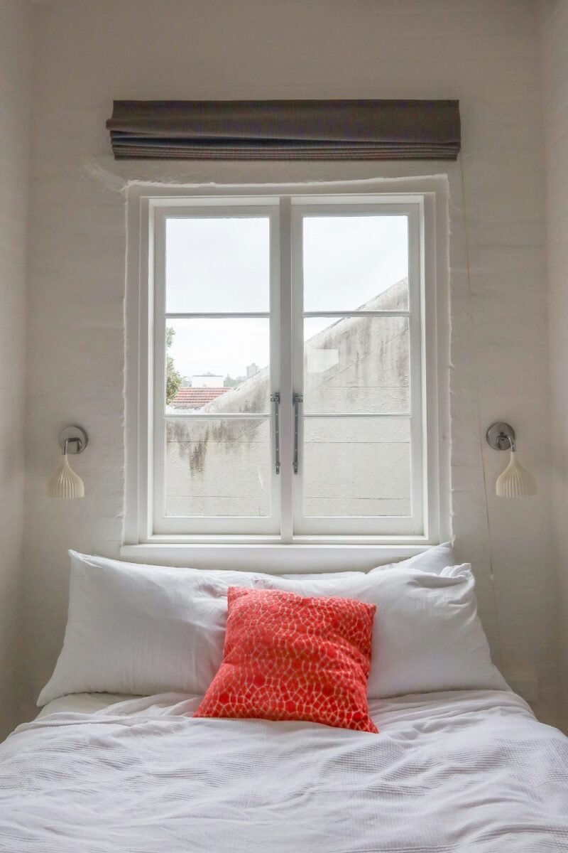 Daylight coming through a white-framed window above a bed in a small bedroom, with a coral cushion and a small wall sconce on each side